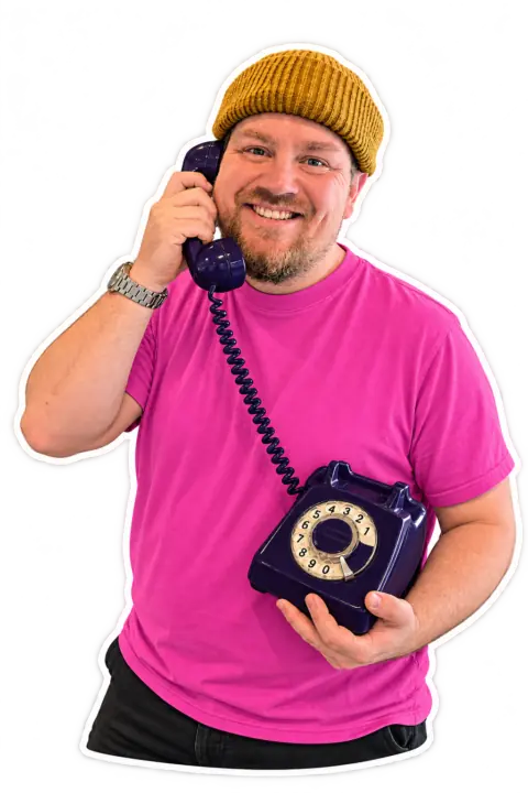 René Kirk i gul hue og pink T-shirt, der taler i en gammeldags mørkeblå telefon med drejeskive